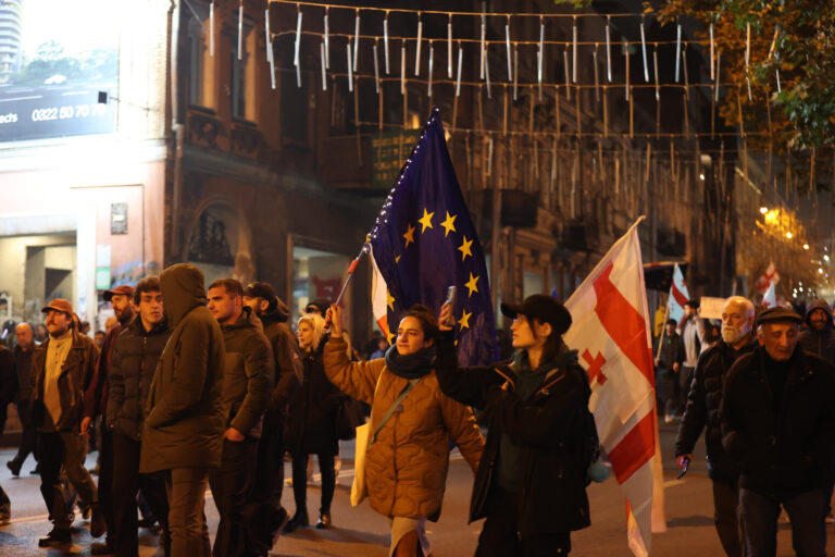 Ein Foto zeigt Menschen mit EU- und Nationalflagge, die bei Nacht durch eine beleuchtete Stadtstraße marschieren.