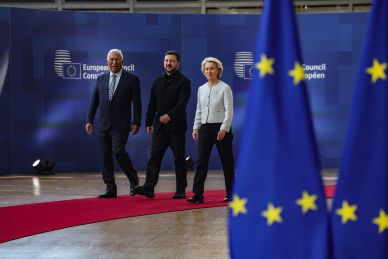 Ein Foto zeigt drei Personen beim Gehen auf einem roten Teppich vor einer blauen Wand mit EU-Logo und EU-Flaggen.